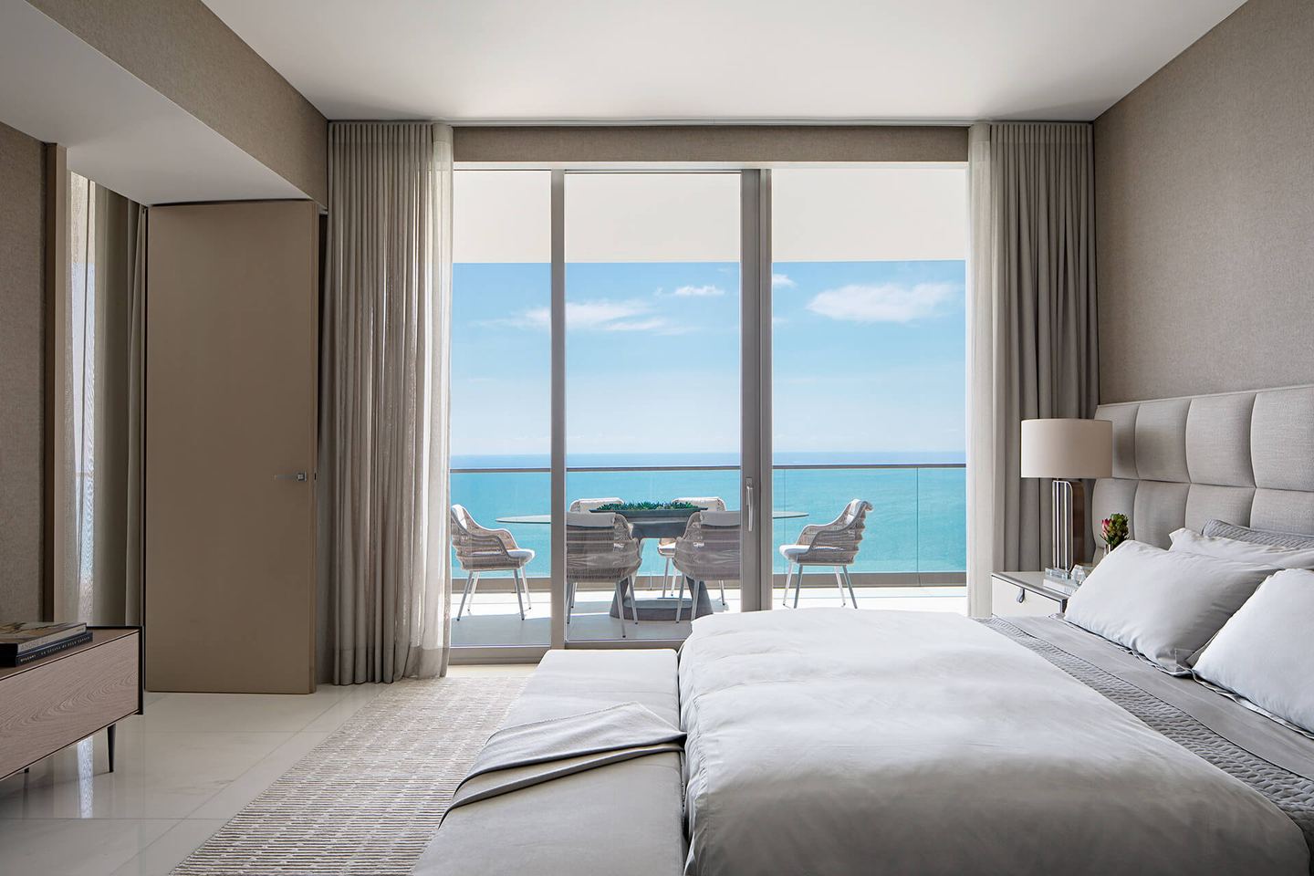 Suíte com interiores em tons neutros, cama ampla e porta de vidro para a varanda com vista para o mar